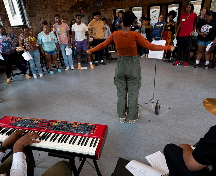 A person spreads their arms at a microphone in front of a crowd. Two musicians sit playing a keyboard and drumset behind her.
