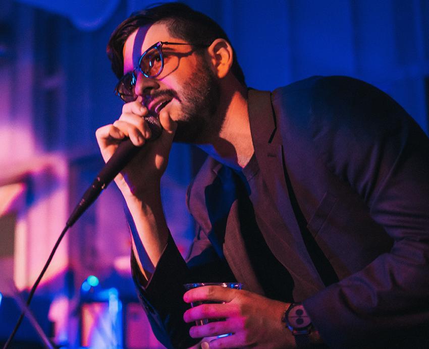 Maggiore talks into a microphone in a club next to a live band