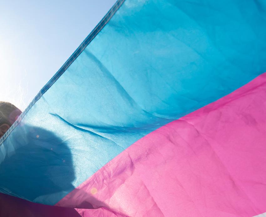 A person in a mask lets a transgender pride flag wave behind them