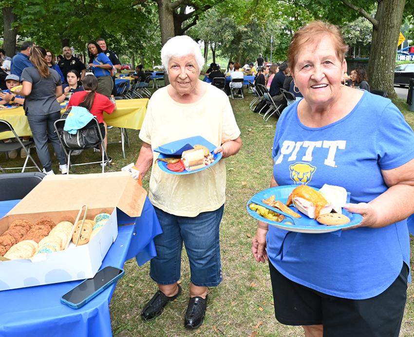 Two people fill paper plates with food. More people eat at outdoor tables in the background