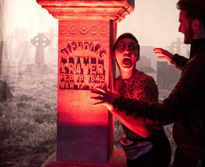 A person holds a gravestone prop and screams for a photo as a person approaches