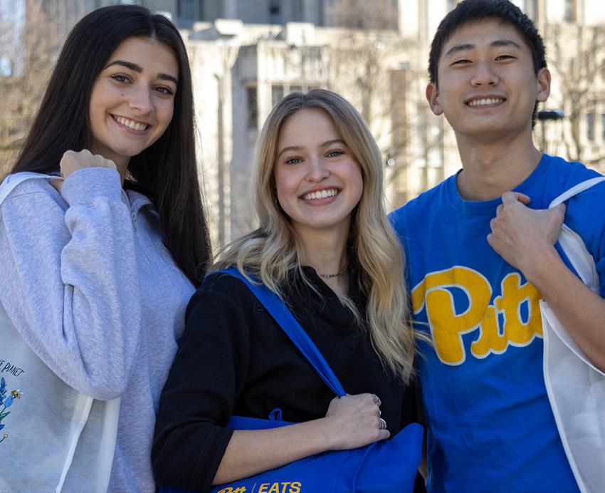 Three students in Pitt gear hold reusable grocery bags on their shoulders