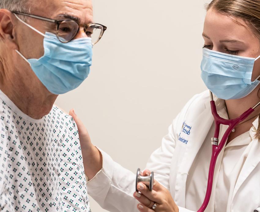 A doctor brings a stethoscope to a patient's chest. Both are wearing face masks