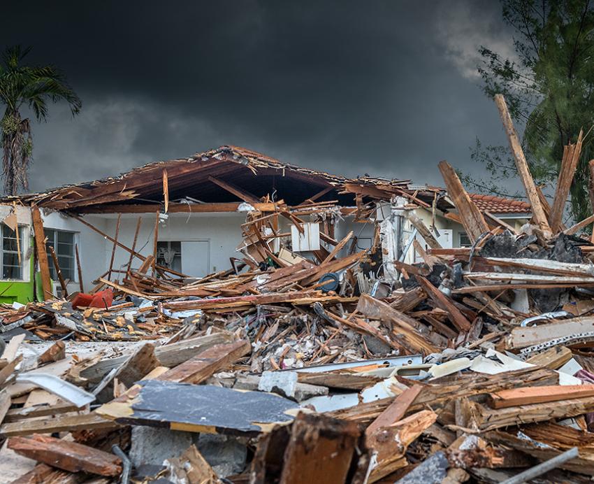 A house is destroyed after a hurricane. The sky is dark and cloudy, and palm trees blow in the wind