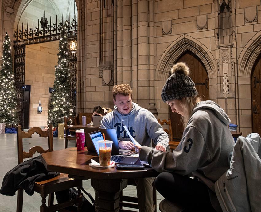 Two students study at a table in the Cathedral of Learning Commons Rooms. Two Christmas trees stand by the entrance.