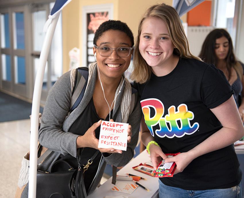 Two students pose smiling. One holds up a hand-written sign that reads Accept Existence or Expect Resistence
