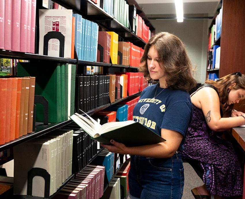 A student reads a book next to a library shelf. Another person writes at a table behind her.