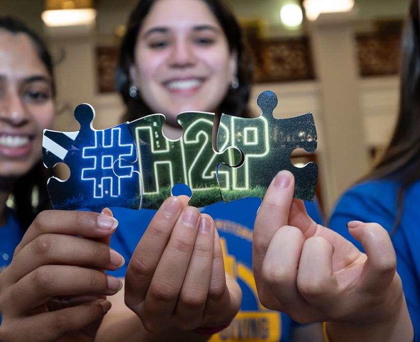 People hold up puzzle pieces to show a neon light that reads (hashtag) H 2 P