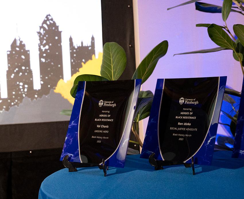 Three plaques sit on a table with a blue tablecloth in front of a projection of Pittsburgh's skyline