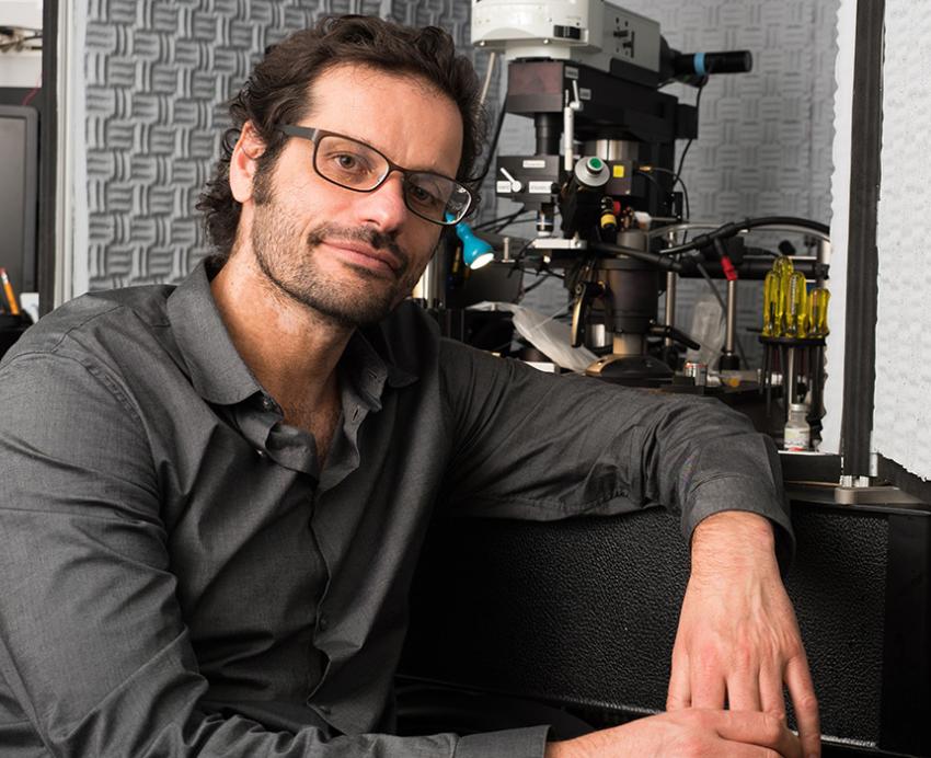 Thanos Tzounopoulos sits on a black bench with a microscope in the background.