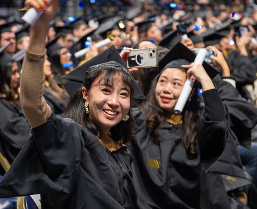 One graduate in regalia waves with her diploma while another takes cellphone pictures