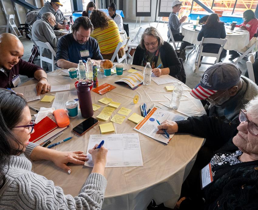 People sit at circular tables, taking notes in small groups