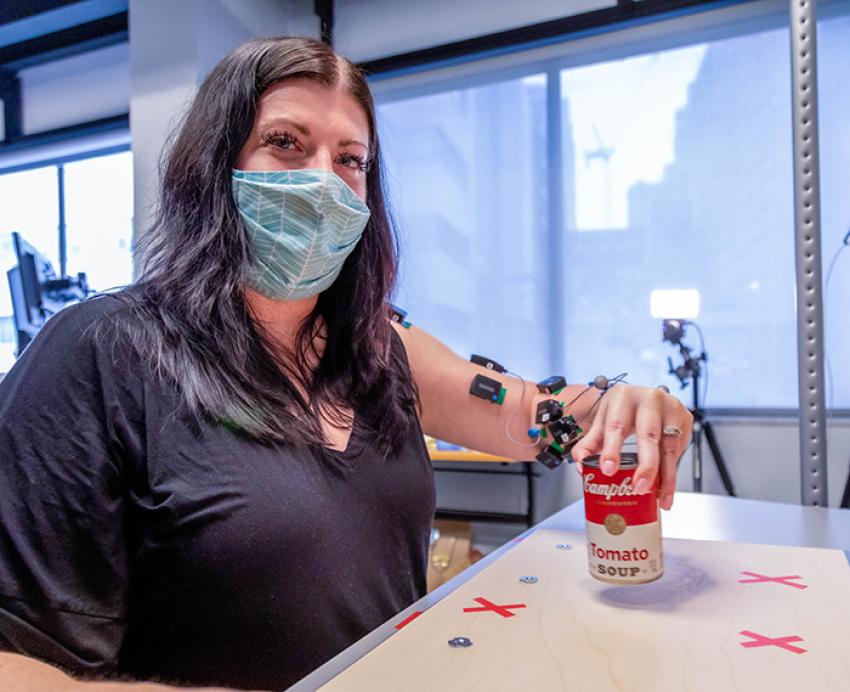 A masked woman lifts a can of soup, aided by devices attached to her left arm