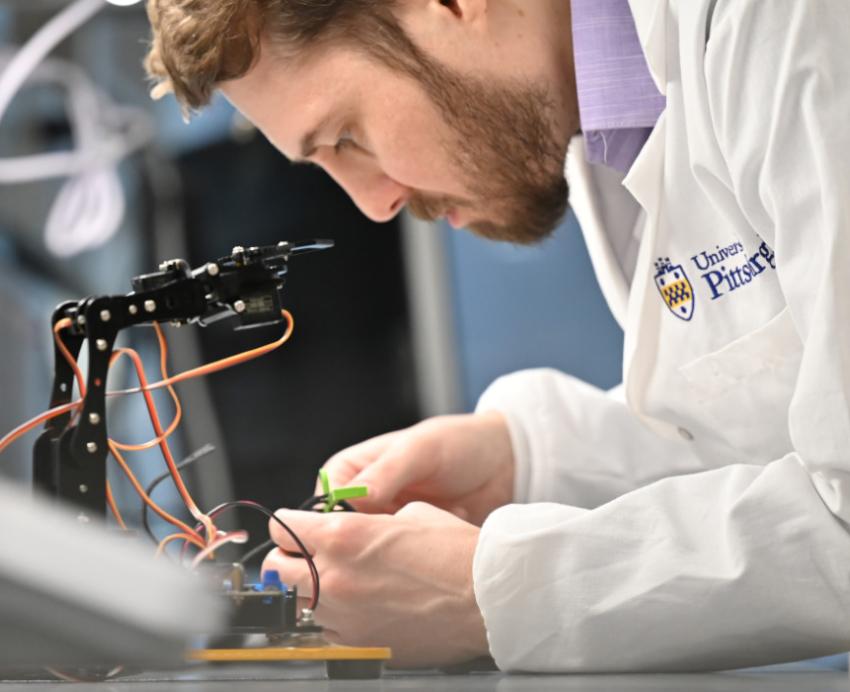 Man wearing Pitt-branded lab coat adjusts wires on electronic device
