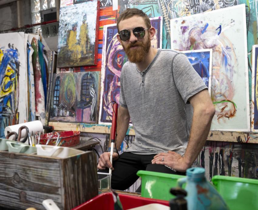 A man wearing glasses, surrounded by abstract paintings.