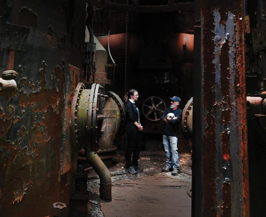 A woman and man stand inside a rusted industrial machine.