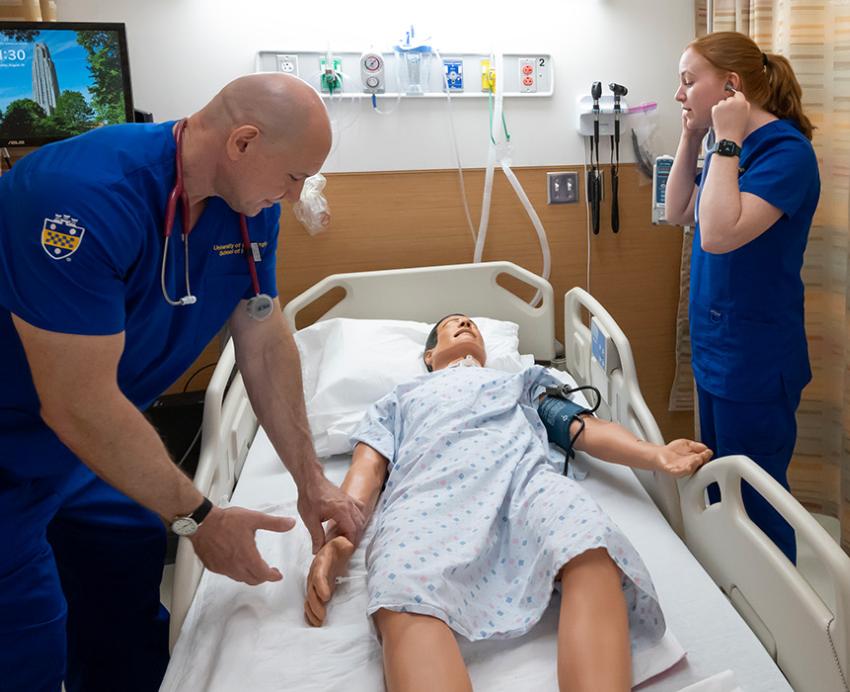 A professor demonstrates taking a pulse on a dummy in a hospital bed