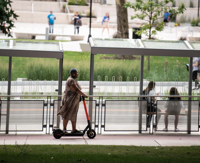 An individual rides an electric scooter on a Bigelow Boulevard sidewalk, passing by a bus shelter.