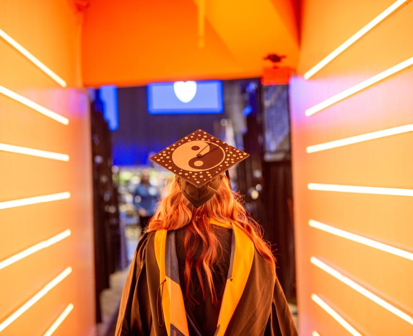 A student stands in a lit hallway in their regalia and personalized cap for Winter Commencement.