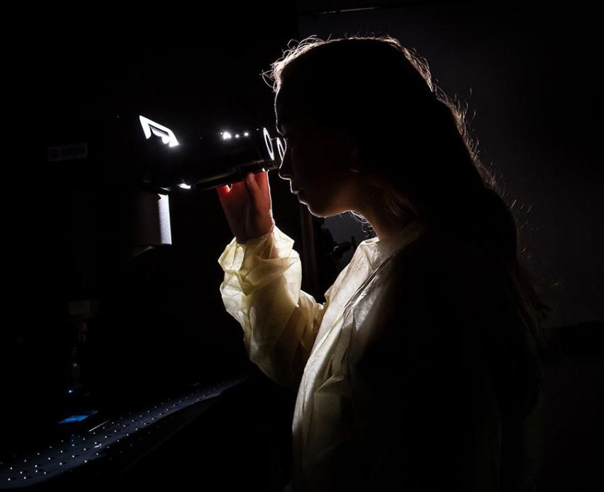 A backlit woman looks into a scope with a computer monitor behind her displays a glowing green image