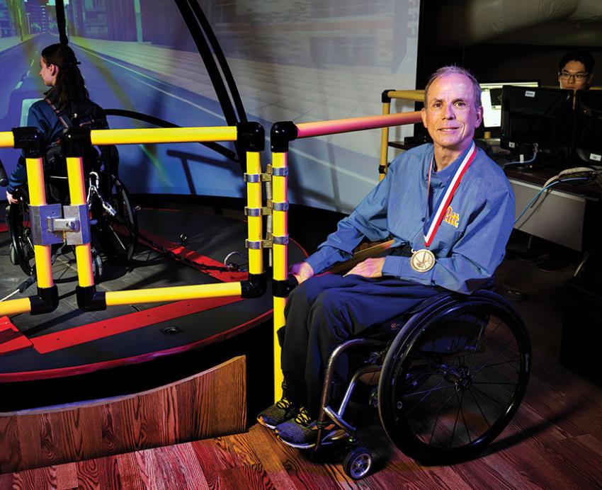 Cooper smiles next to a simulation device, which a person in a wheelchair is using. Cooper is wearing a medal.