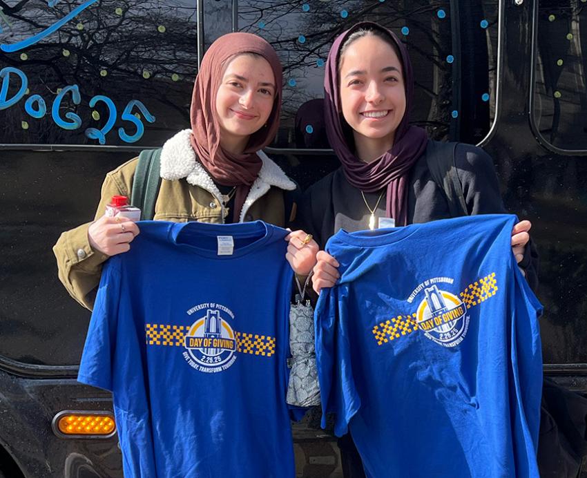 Two people hold up Pitt Day of Giving shirts in front of a black shuttle bus