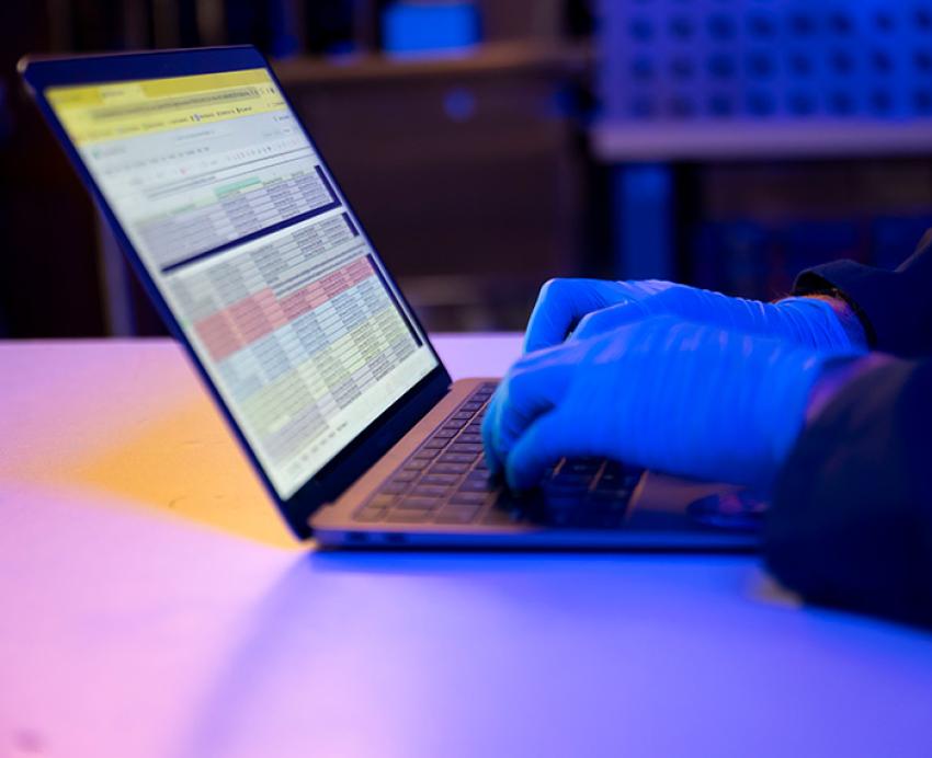 Gloved hands typing on a laptop, lit in blue and purple