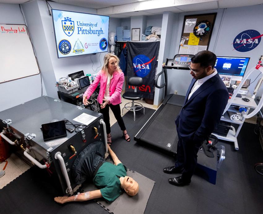 Two people look down at a mannequin laying in a machine.