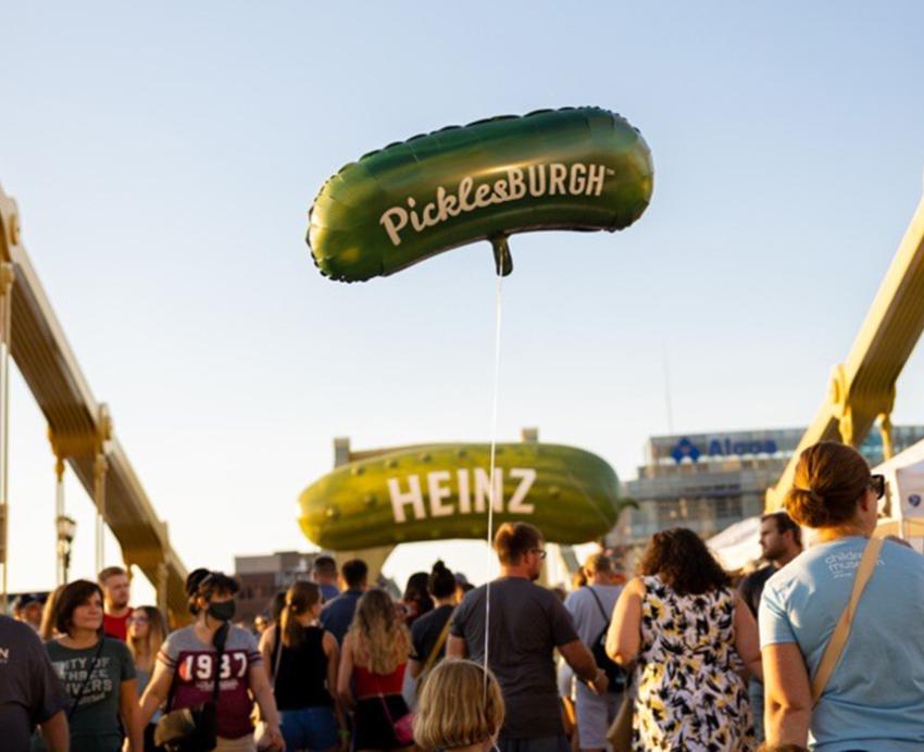 Many people on one of the Pittsburgh bridges, a child carries a pickle-shaped balloon that reads "PicklesBURGH".  In the distance there is a pickle-shaped sign reading "HEINZ".