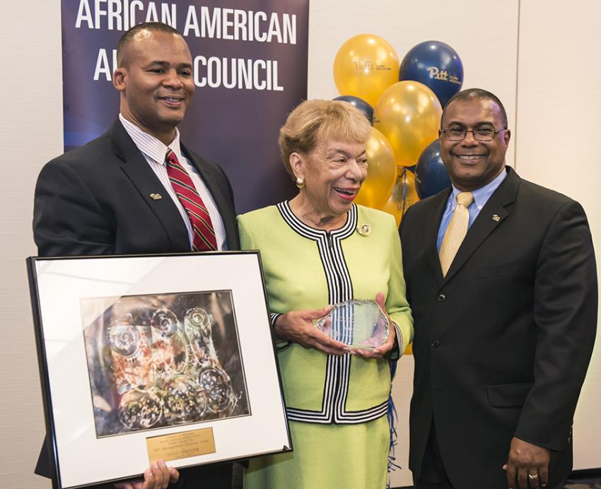 A woman receives an award while standing next two two men in front of a sign that reads "African American Alumni Council"