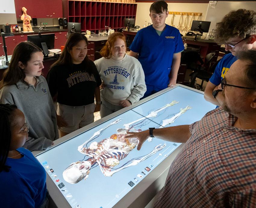 A professor gestures to a digital anatomy table, which students congregate around.