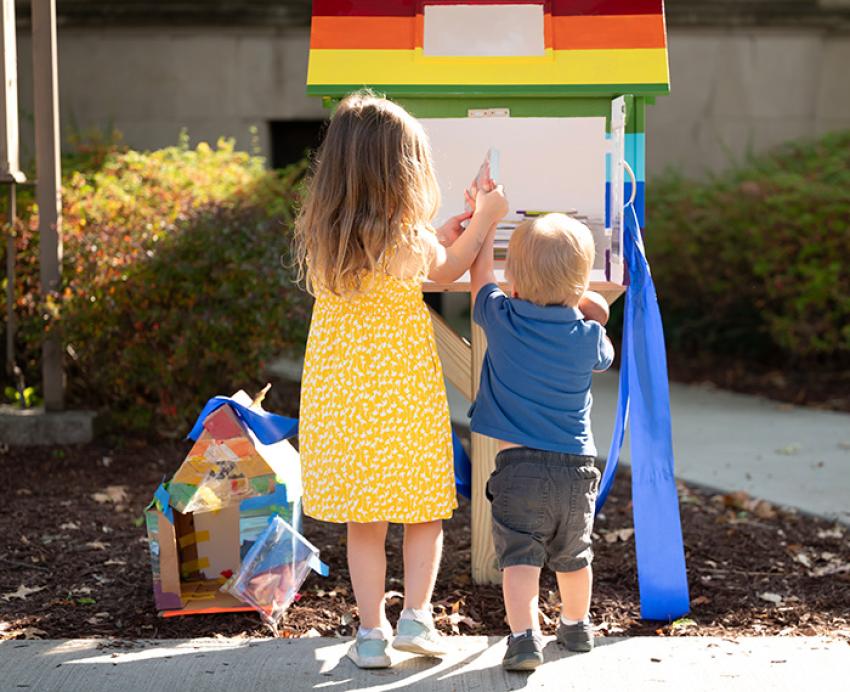 Two children place books in a Little Free Library painted in rainbow colors. A prototype of the box sits on the ground nearby.