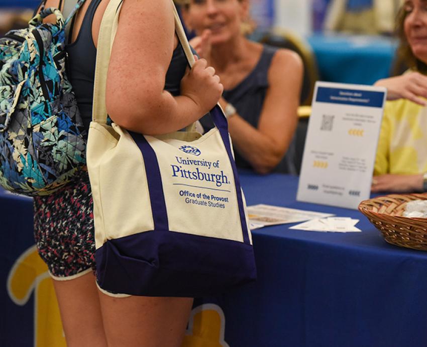 Person carries University of Pittsburgh tote bag while talking to women at a booth