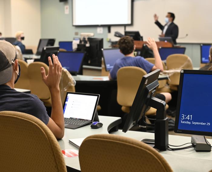 Students in masks in computer classroom, raising hands as teacher lectures