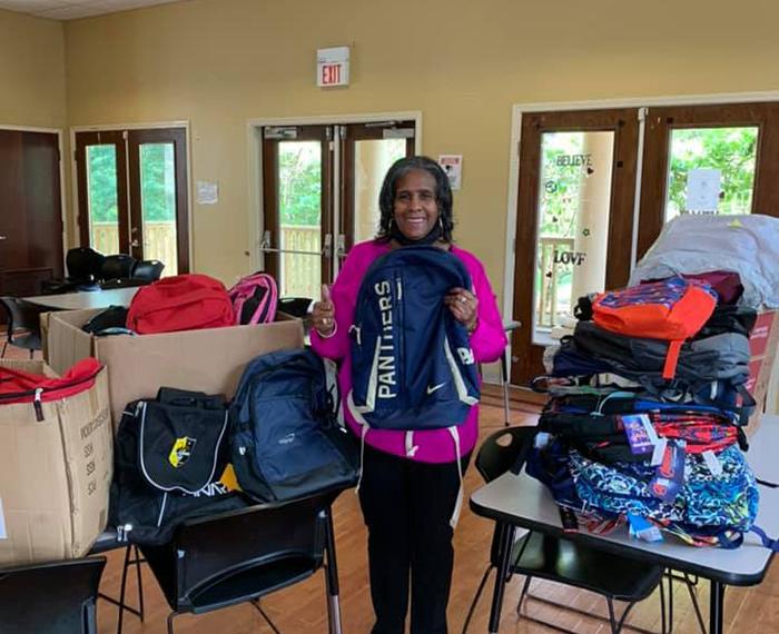 Woman with purple shirt holding thumbs up next to piles of backpacks