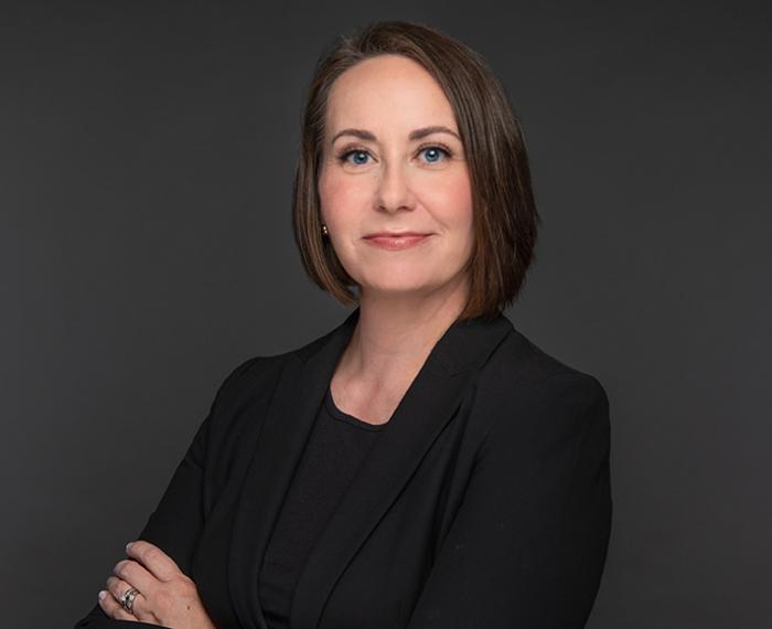 Lina Dostilio in black suit with arms folded in front of grey background