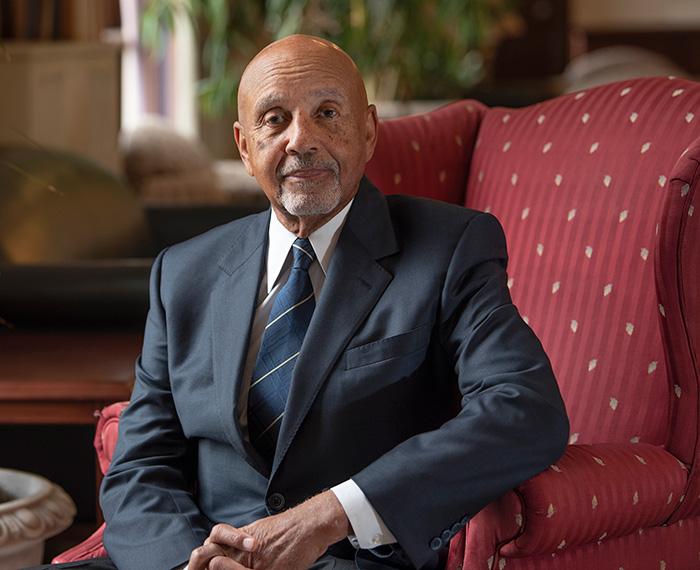 Jones sits in a red chair wearing a dark grey suit
