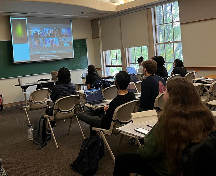 Students in rows of desks watch a video call on a projector