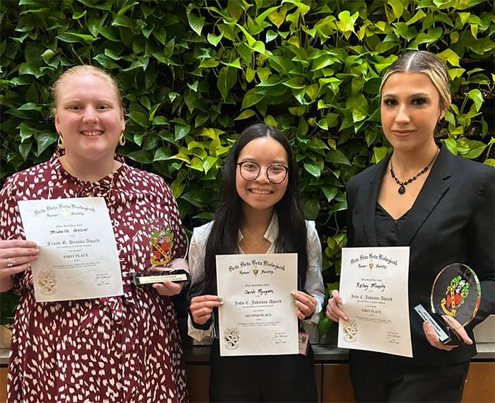 A person in a red and white dress, a person in a white shirt and a person in a black suit hold up certificates and awards