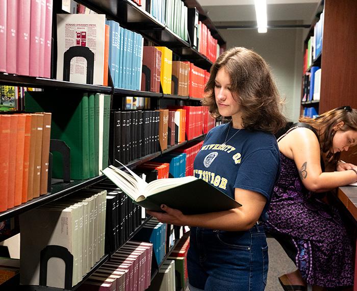 A student reads a book next to a library shelf. Another person writes at a table behind her.