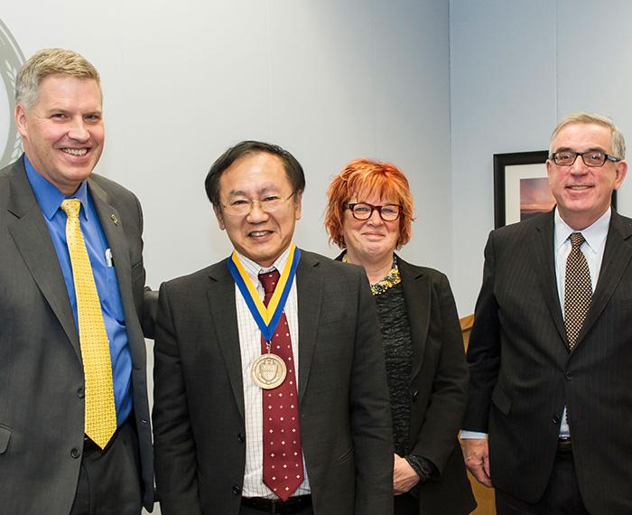 Four people in suits, one in the middle wears a medal around his neck