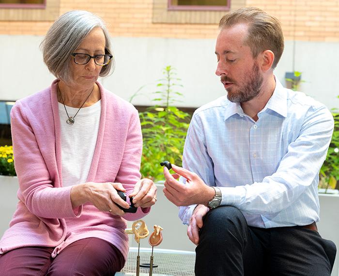 Two people hold small black hearing aids