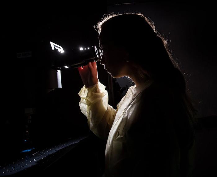 A backlit woman looks into a scope with a computer monitor behind her displays a glowing green image
