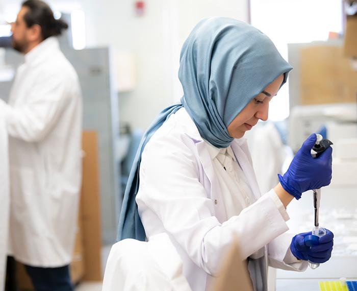 A researcher in a white coat and hijab fills a tube with a pipette.
