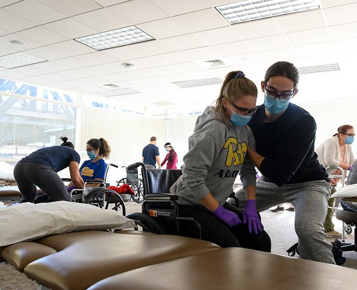 Physical therapists help two people out of wheelchairs in a clinic. Everyone is wearing face masks.