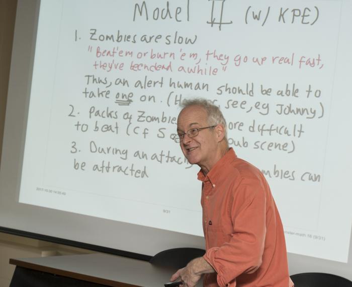 Ermentrout in an orange shirt in front of a screen with some rules of the math lesson on it