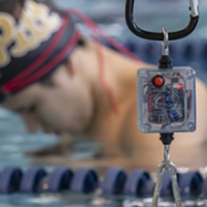 An electronic device hanging by a clip with a swimmer with a Pitt cap on in the background