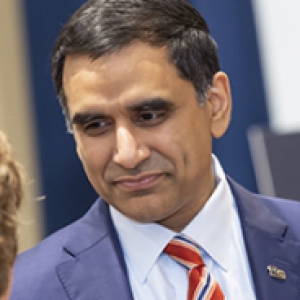 A man in a blue suit with a small pin on his lapel and a red striped tie speaks with another person