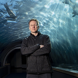 Clark, wearing a dark pullover and pants, standing in an aquarium tunnel with sea life swimming behind him