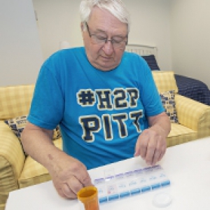 Dennis Stark, in light blue Pitt T-shirt sitting in a plaid yellow armchair, placing pills into a pill counter placed on a white table. A prescription pill bottle is on the table.
